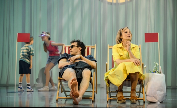 Två barn står i bakgrunden medan en man i solglasögon och en kvinna i gul klänning sitter i solstolar.