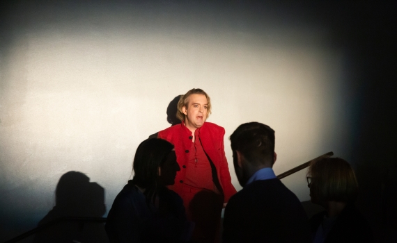 Andreas T Olsson som Dante i ett mörkt rum, han lutar sig mot väggen och man kan ana siluetter av människor runt honom.