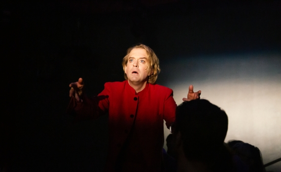 Andreas T Olsson som Dante i ett mörkt rum där man bara kan ana siluetter av människor runt honom.