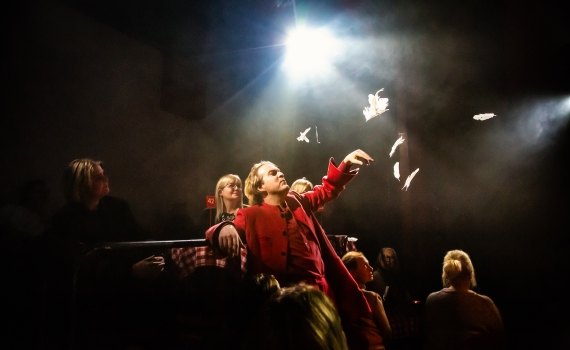 Andreas T Olsson som Dante, han lutar sig mot ett räcke och kastar ut fjädrar i luften. Rummet är fullt av människor. 