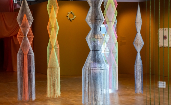 Flera höga, geometriska skulpturer av färgade trådar hänger i ett rum med senapsgula väggar och trägolv.
