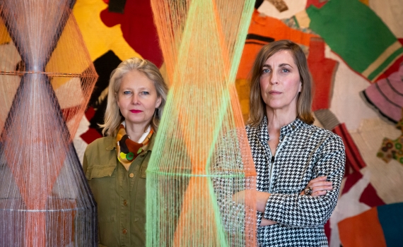 Två kvinnor står bland färgade trådskulpturer, med ett textilt konstverk i bakgrunden.