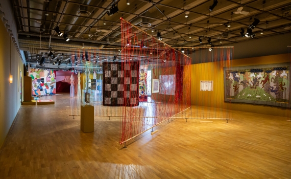 Överblick över ett galleri med trådbaserade installationer, väggbonader och färgstarka textilverk.
