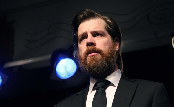 En man i svart kostym och svart slips som läser från en bok