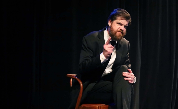 En man i svart kostym och svart slips som sitter på en stol och grimaserar