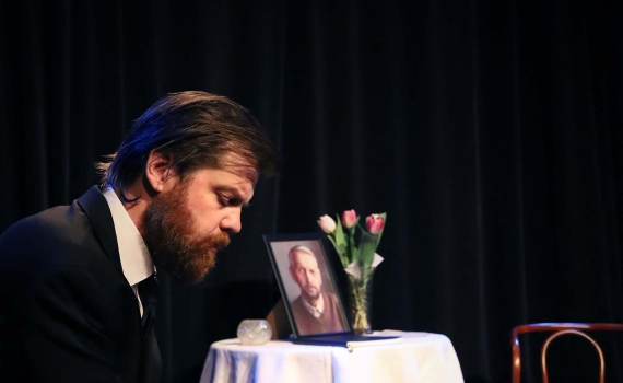 En man i svart kostym och svart slips försjunken i tankar, i bakgrunden syns ett bord med ett foto och en vas blommor