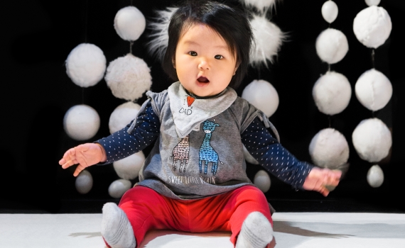 Spädbarn med svart hår sitter på golvet, klädd i grå tröja, röda byxor och haklapp.