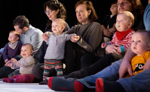Vuxna och spädbarn sitter på golvet i publik, flera barn hålls i famn eller står med stöd.