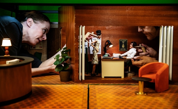 Kvinna i svart tröja styr en docka i miniatyrrum, en annan docka manipuleras av hand från höger.