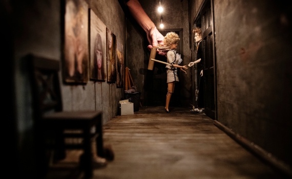 Dockor i en mörk korridor med målningar på väggen, två händer manipulerar figurerna.