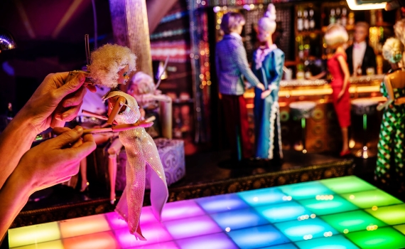 Docka i glittrig klänning hålls ovanför upplyst dansgolv, fler dockor står vid bardisk i bakgrunden.