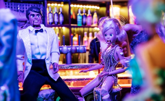 Dockor vid en bardisk i klubbmiljö, en docka i paljettklänning ser överraskad ut.