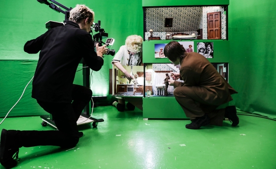 Tre vuxna personer arbetar med dockor och miniatyrmiljö i en green screen-studio, en person filmar.