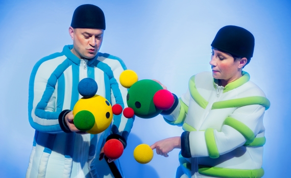 Två vuxna i färgglada dräkter håller mjuka, färgglada bollar med ansikten och antenner.