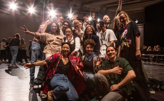 Grupp på ca 18 unga vuxna poserar glatt på en scen, flera gör segertecken och ler mot kameran.