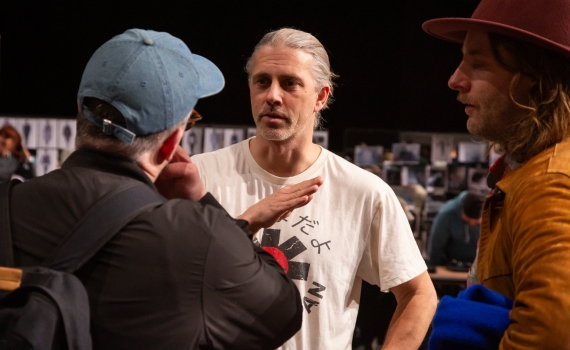 Tre män i samtal, en med vitt hår i hästsvans bär vit t-shirt med tryck, de andra har keps respektive hatt.