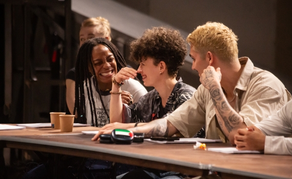 Tre unga vuxna sitter vid ett bord och skrattar, en kvinna med rastaflätor, en med lockigt hår och en tatuerad man.