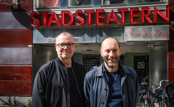 Två medelålders män står utanför Stadsteatern, båda bär mörka jackor och glasögon.