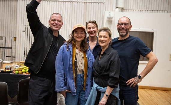 Fem vuxna, tre kvinnor och två män, poserar i en replokal, fruktfat syns i bakgrunden.