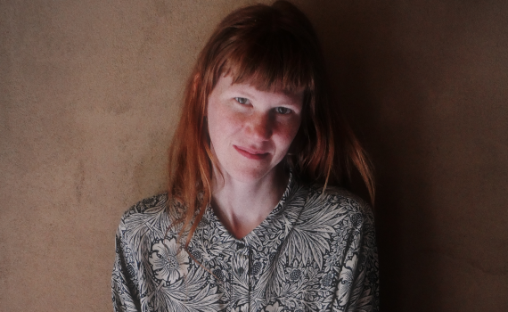 Ung kvinna med långt rött hår och lugg, bär mönstrad skjorta, står framför brun vägg och ler svagt.