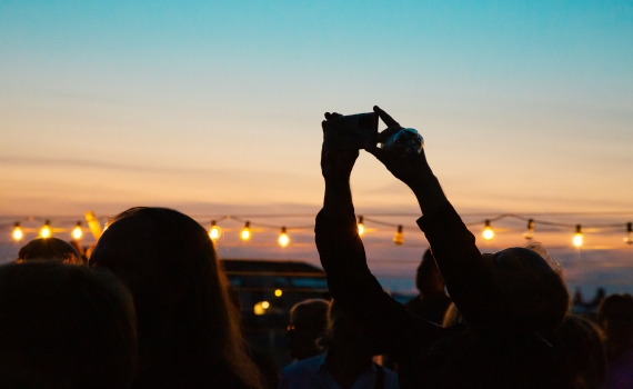 Silhuetter av personer i motljus vid solnedgång, en person håller upp en mobil mot himlen.