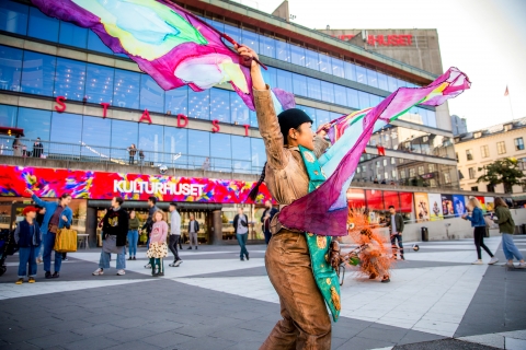 En skådespelare från Marionetteatern håller i stora färgglada flaggor på Sergels torg. 