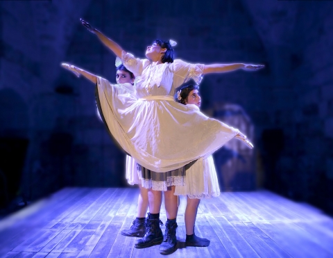 Tre personer i vita klänningar, där en hoppar högt i ett danssteg. 