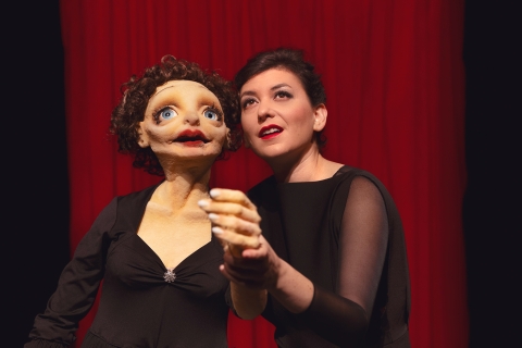 En kvinna i svart klänning håller i en stor docka som påminner om Edith Piaf i svart klänning. 