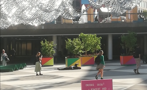 Man ligger på rygg på Sergels torg och spelar trumpet när människor passerar.