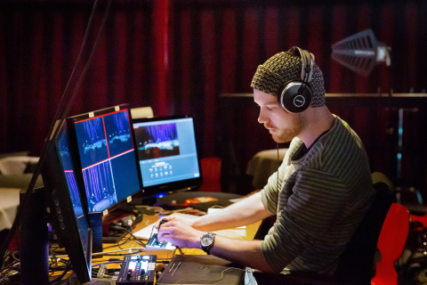 En man med hörlurar sitter vid ett bord med flera datorskärmar och reglage på. 