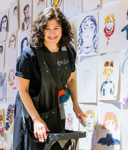 En leende kvinna med förkläde med målarfärg på står på en stege och sätter upp teckningar och målningar på en stor vägg.