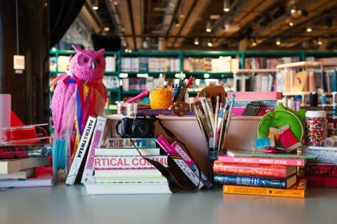 Bord med konstmaterial som penslar, garn, kamera och staplade böcker i färgglada omslag.