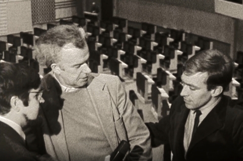 Svartvit bild av tre män i samtal i en tom biosalong eller teater, två i kavaj och en i glasögon. En av männen håller en mikrofon.