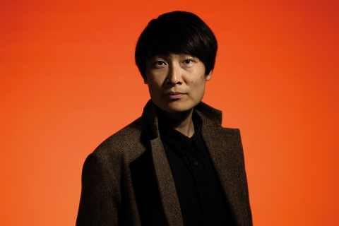 Person med kort, mörkt hår i brun kavaj och svart skjorta, poserar mot orange bakgrund.