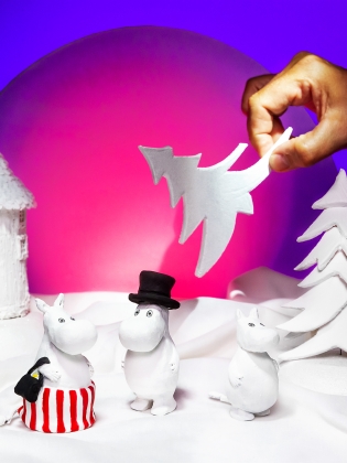 Tre mumintroll i ett vintrigt landskap, en människohand håller i en vit gran.