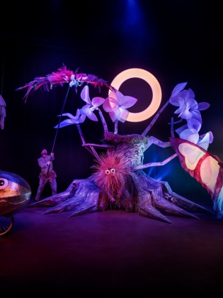 En skådespelare med fjärilsvingar, en med en cykel i form av en fisk, mitt i allt står ett träd. 