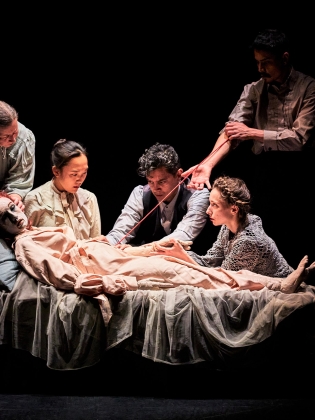 En stor docka ligger ner på en säng omgiven av fem personer.