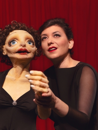 En kvinna i svart klänning håller i en stor docka som påminner om Edith Piaf i svart klänning. 