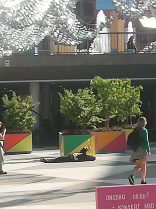 Man ligger på rygg på Sergels torg och spelar trumpet när människor passerar.