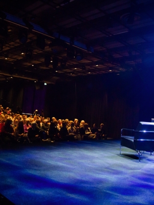 En publik sitter och väntar framför en scen, på scenen står det två tomma stolar. 