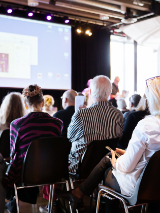 En publik sitter på en konferens framför en scen. 