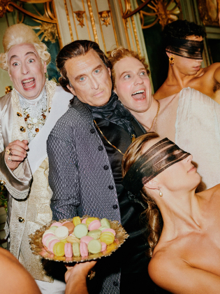 En excentrisk scen med personer klädda i historiska kläder, där några bär ögonbindlar och andra ser chockerade eller glada ut. Färgglada macarons serveras på ett fat.