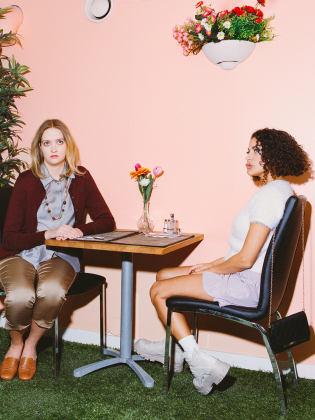 Två kvinnor sitter mittemot varandra vid ett bord i ett pastellfärgat rum med konstgjord gräsmatta och blommor på väggen.