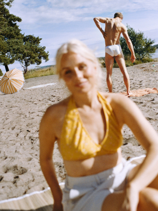 Kvinna i gult bikinilinne sitter suddigt i förgrunden på en sandstrand, medan en man i vita badkläder står i bakgrunden vid vattnet.