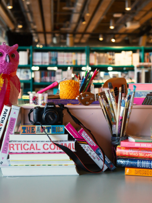 Bord med konstmaterial som penslar, garn, kamera och staplade böcker i färgglada omslag.