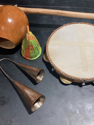 Fyra musikinstrument på en mörk yta: en berimbau, en caxixi, en tamburin och två agogô-klockor.