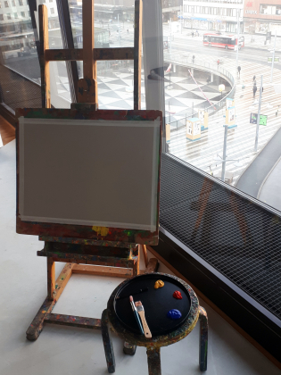 Staffli med en tom duk och en målarpall med tre färgdroppar och penslar, vid ett fönster med utsikt över en stadsmiljö.