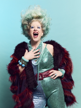 Kvinna i stort hår, turkos glittrig byxdress och mörkröd pälsjacka sjunger.