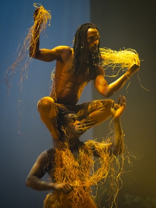 En man står hukad på en annan mans axlar. De båda håller i halm.