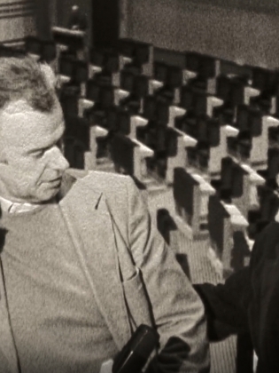 Svartvit bild av tre män i samtal i en tom biosalong eller teater, två i kavaj och en i glasögon. En av männen håller en mikrofon.